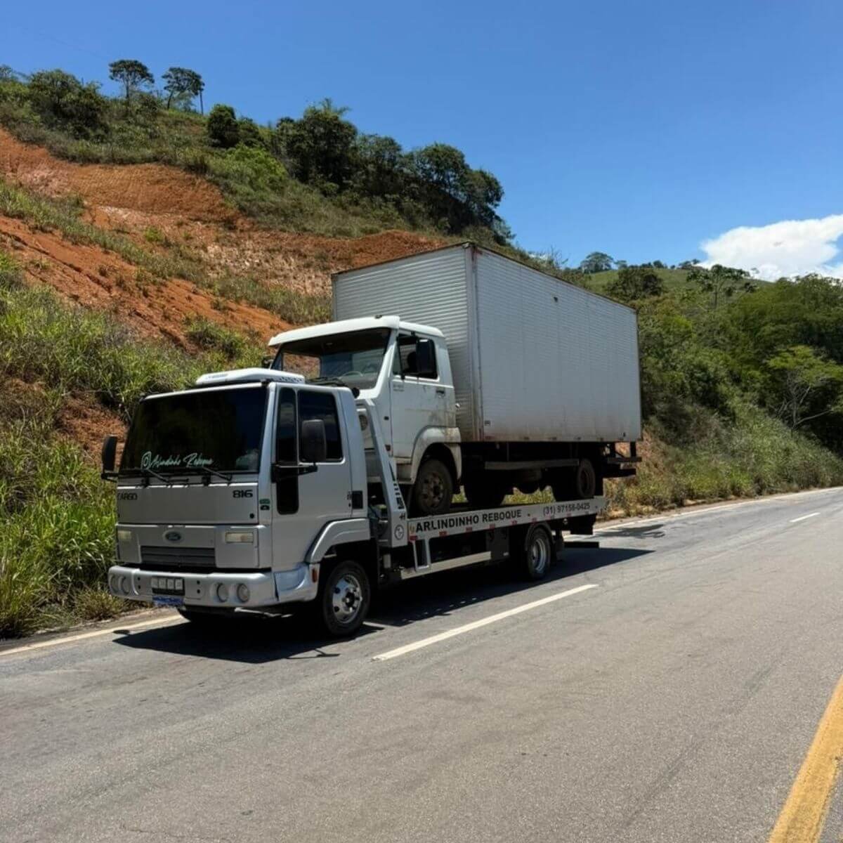 Guincho levando caminhão baú branco em rodovia com vegetação ao fundo.