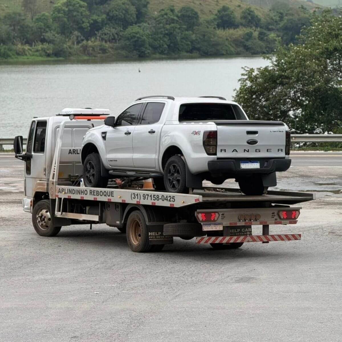 Guincho transportando picape branca próximo a uma lagoa/represa.